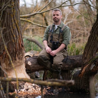 Person wearing chest waders sits on a fallen tree in a wetland area beside trees showing evidence of beaver activity.